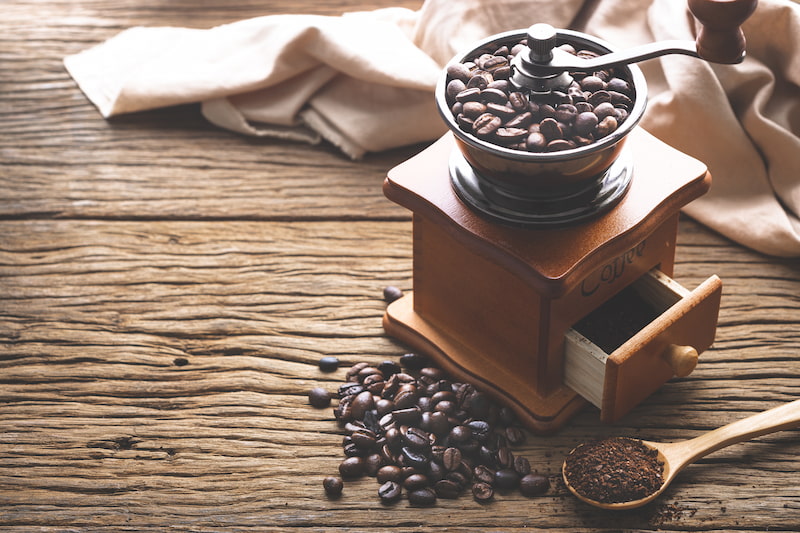 a manual coffee grinder with coffee beans inside, a small drawer with ground coffee, loose beans on the table and a wooden spoon with ground coffee on a rustic wooden surface.