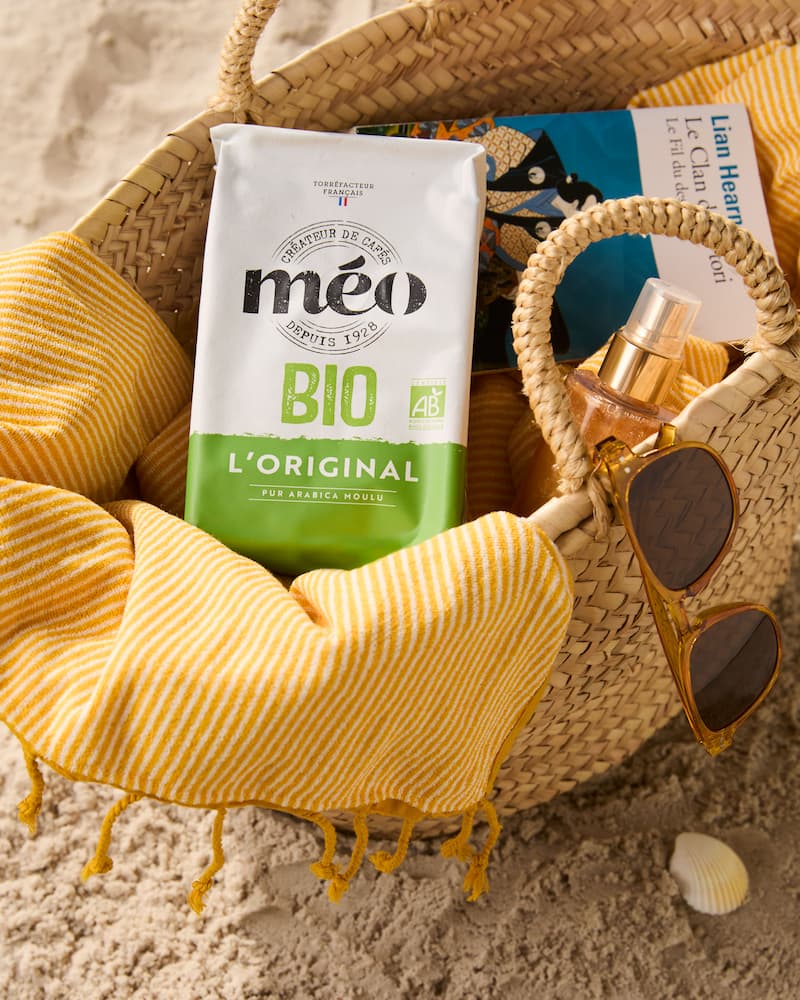 straw bag on the sand with a yellow striped towel, sunglasses, a spray can, a book, a seashell and a bag of méo bio organic coffee nearby.