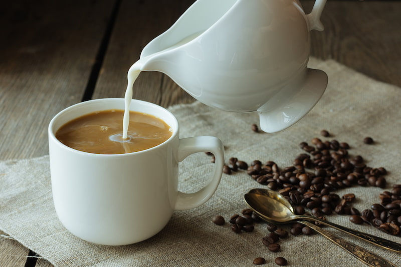 milk is poured into a white coffee cup on a canvas, surrounded by coffee beans and two spoons.