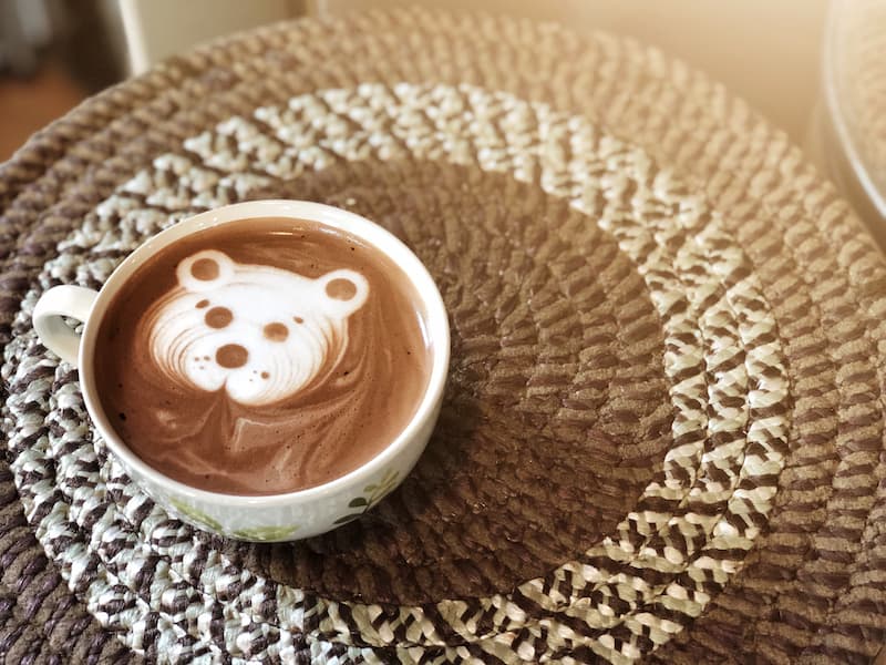 a cup of coffee with foam resembling a bear's face sits on a circular woven placemat.
