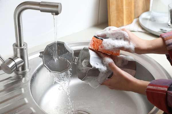 person washing a coffee pot with a sponge under running water in the kitchen sink.