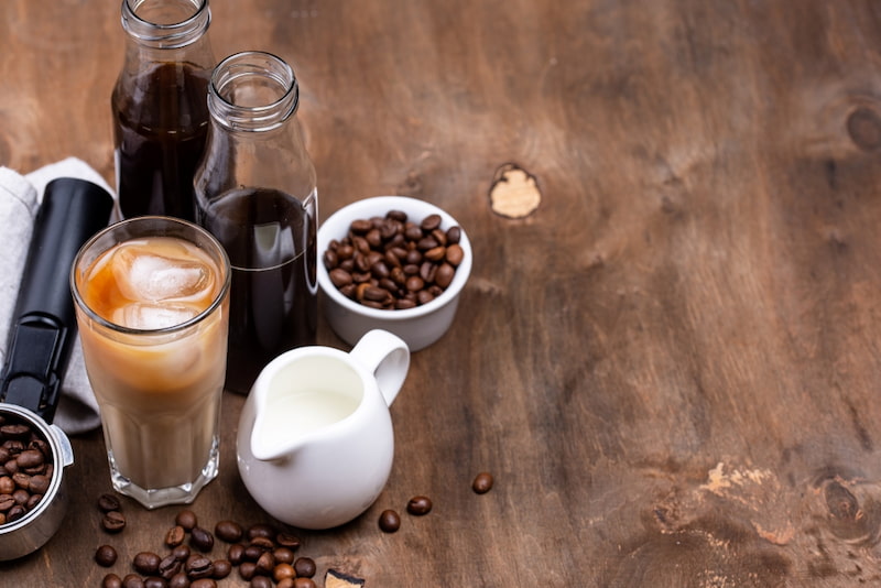 cold coffee in a glass with milk, two bottles of cold coffee, a small jug of milk, coffee beans in bowls and a coffee press on a wooden table.