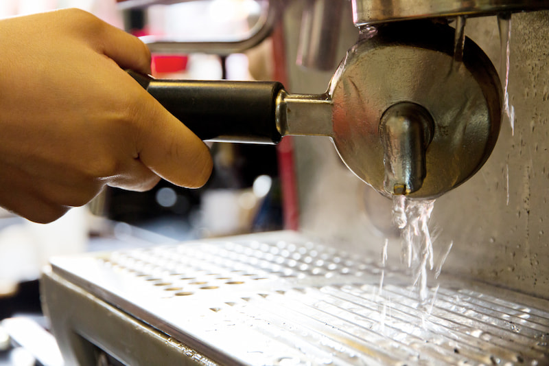 hand holding a portafilter under the running water of a coffee machine, with water splashing in the drip tray.
