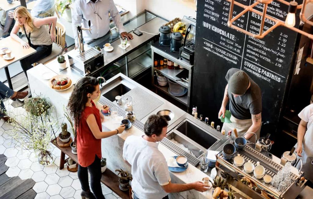 people inside a crowded bar with bartenders preparing drinks behind the counter, while customers wait and sit at tables. a tab menu is visible in the background.