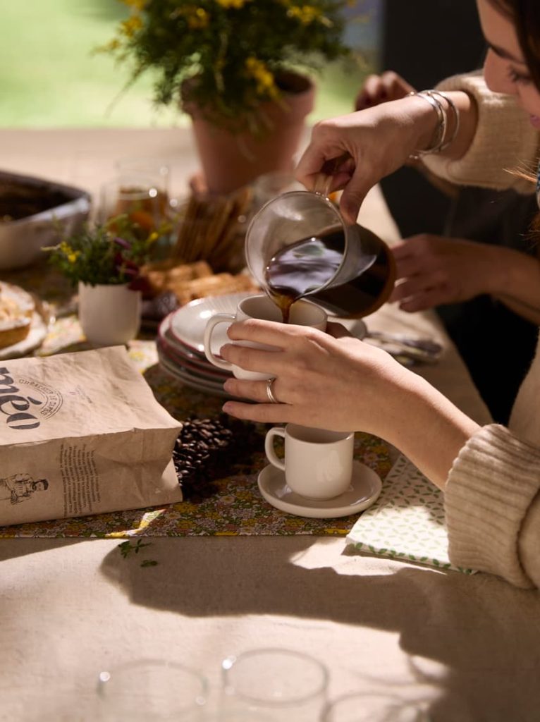 A person pours coffee from a glass carafe into a white cup placed on a table set with food, a bag of coffee beans and a potted plant.