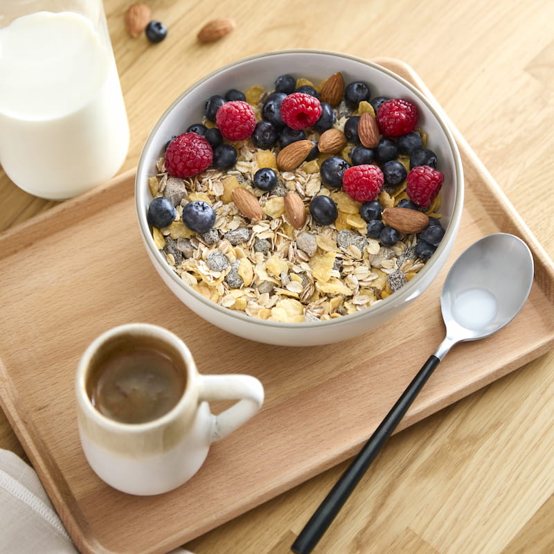 a bowl of granola with berries and almonds, a glass of milk and a cup of espresso on a wooden tray with a spoon.
