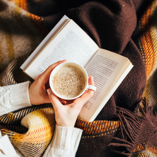 person holding a cup of coffee and reading a book wrapped in a checkered blanket.