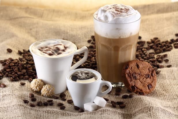 three types of coffee drinks with whipped cream and chocolate chips. next to them on a canvas surface a biscuit, coffee beans and sugar cubes are placed.