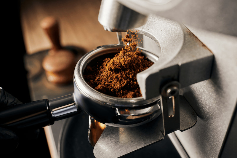 close-up of ground coffee poured into a filter holder from a coffee grinder. a pestle is blurred in the background on a wooden surface.