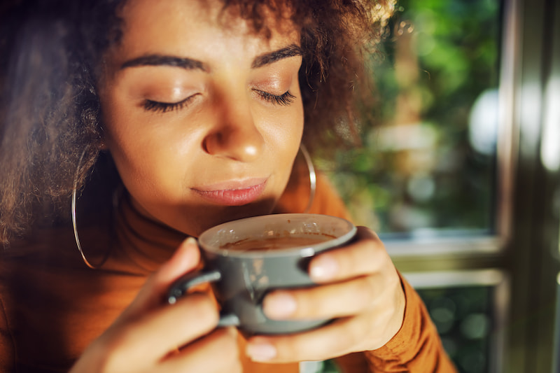 a woman with her eyes closed holds a cup close to her face, appearing to enjoy the aroma. She has curly hair and wears hoop earrings and a brown top. the background shows greenery and a window.