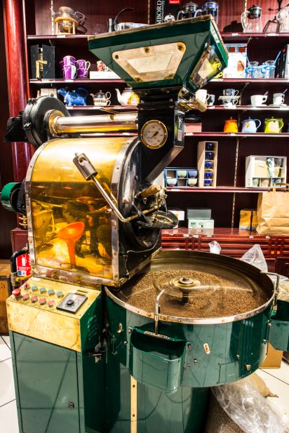 a green and gold coffee roasting machine with control panel in a shop window, surrounded by various teapots and cups on shelves in the background.