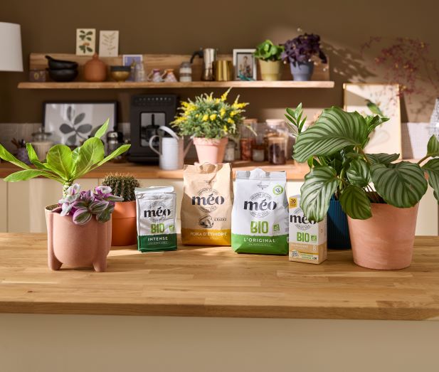 Four bags of Méo coffee and a carton of Méo coffee pods are displayed on a wooden worktop surrounded by various potted plants in a kitchen.
