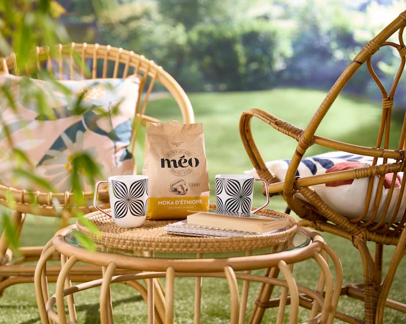 a rattan patio set with two cups, a meo coffee bag and a notebook sits on a glass table in a sunny garden.
