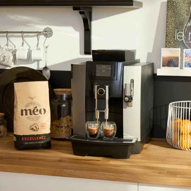 a modern espresso machine brews coffee in two transparent glasses on a wooden counter, next to a bag of coffee beans and a wire fruit basket with oranges. in the background there are shelves with cups.