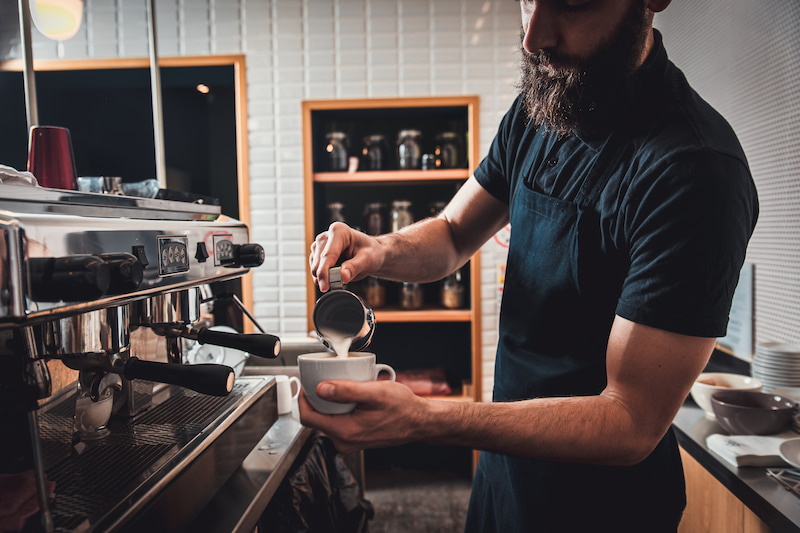 bearded barista pouring milk into a cup of coffee from a metal jug next to an espresso machine in a cafe.