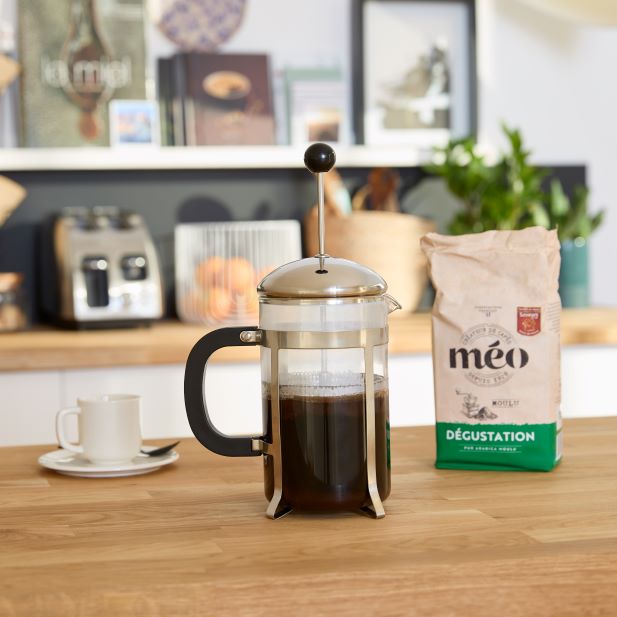 A French press full of coffee sits on a wooden kitchen counter, next to a bag of meo coffee beans. A white coffee cup and saucer are positioned in the background.