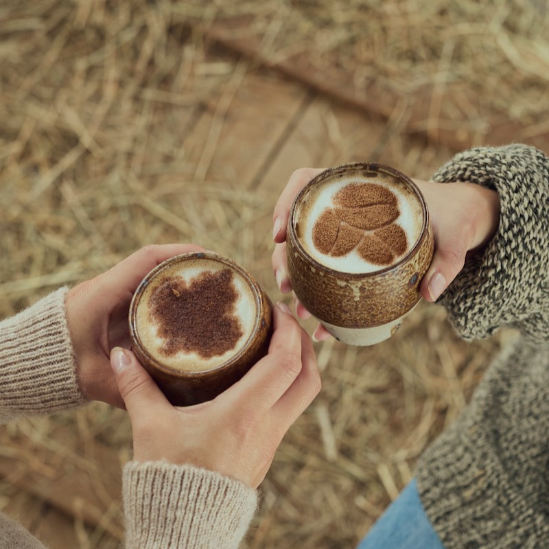 two people holding cups of cappuccino with cocoa powder designs on the foam, one resembling a bear and the other a leaf, on a straw-covered floor.