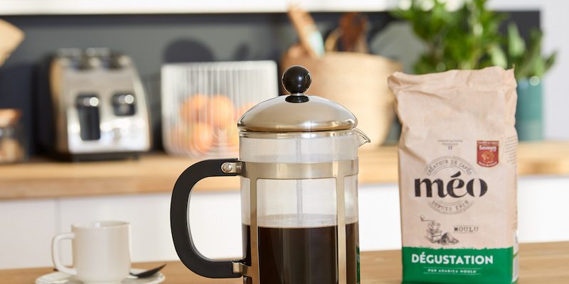 A coffee pot full of coffee sits on a wooden kitchen counter next to a bag of meo-tasting coffee, with a cup and saucer in the background.
