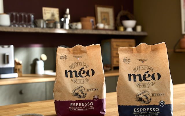 Two bags of méo espresso coffee are displayed on a wooden counter, with a cup of coffee and waffle biscuits in the foreground in a modern kitchen.