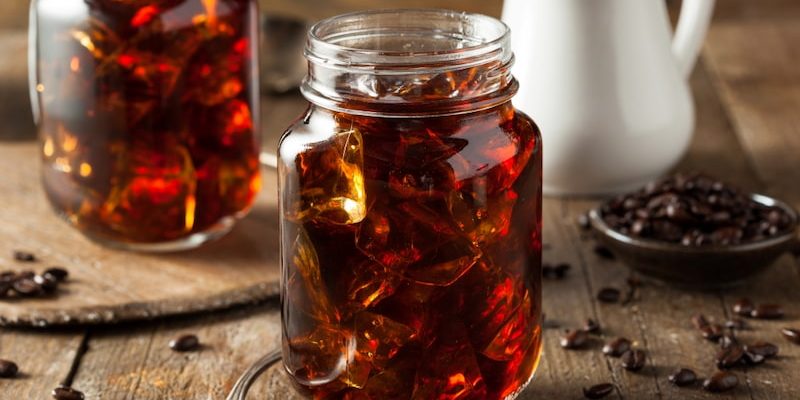 two glass jars full of cold coffee lie on a wooden table next to a spoon, coffee beans and a small jug.