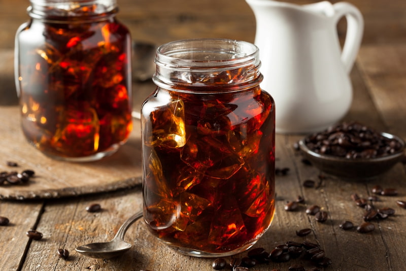 two glass jars full of cold coffee lie on a wooden table next to a spoon, coffee beans and a small jug.