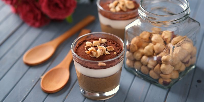 two glasses of layered chocolate dessert garnished with nuts sit on a blue wooden table next to two wooden spoons and a jar of hazelnuts with red flowers in the background.