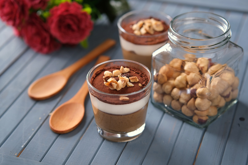 two glasses of layered chocolate dessert garnished with nuts sit on a blue wooden table next to two wooden spoons and a jar of hazelnuts with red flowers in the background.