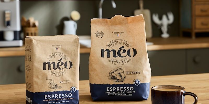 two bags of méo espresso coffee, one ground and the other in beans, are placed on the wooden kitchen counter, next to a small blue cup, a saucer and a spoon.