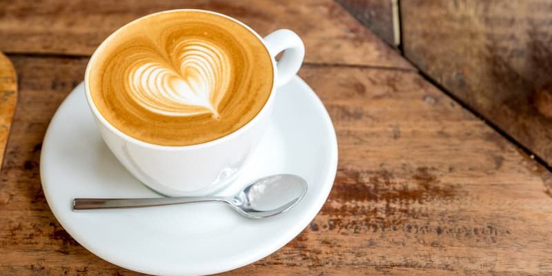 on a wooden table, a cup of coffee with heart-shaped latte art is placed on a white saucer with a metal spoon.