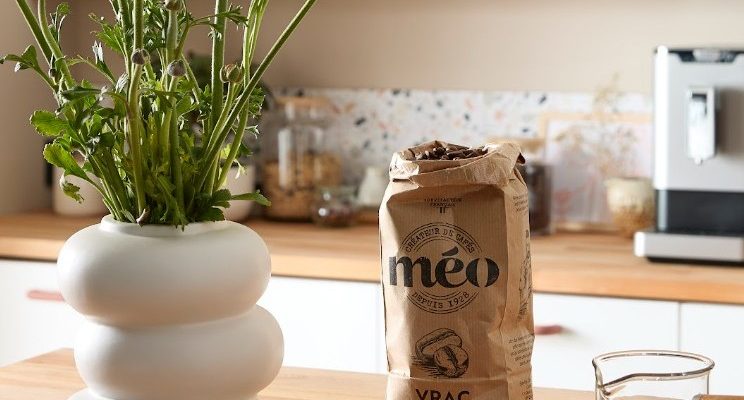 a bag of meo coffee, a white vase with yellow flowers, a glass coffee carafe and a cup are placed on a wooden kitchen counter with shelves and a coffee machine in the background.
