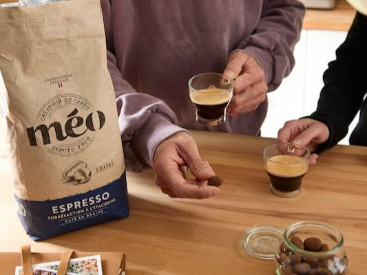 two people stand in front of a wooden table holding espresso in glass cups; nearby are a bag of méo espresso and a jar of chocolate-covered almonds.