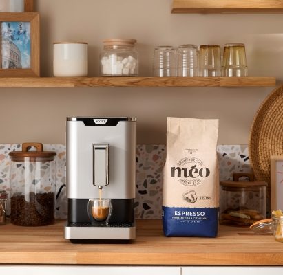 There's a silver coffee machine on the kitchen counter, next to a bag of Meo espresso. on the rear wall you can see shelves with jars, glasses, photos and decorations.