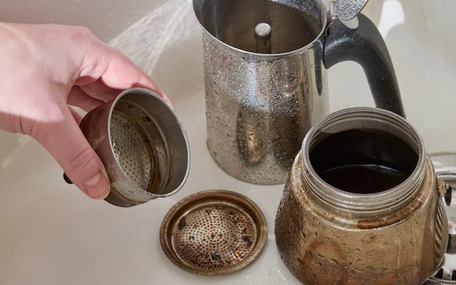 a disassembled moka pot in a sink, with visible coffee residue. a hand holds one part, while other components are scattered nearby, including a filter and a gasket.