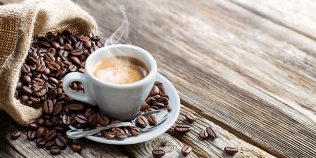 a white cup of steaming coffee sits on a saucer with a spoon, surrounded by scattered coffee beans spilling out of a burlap sack onto a wooden surface.