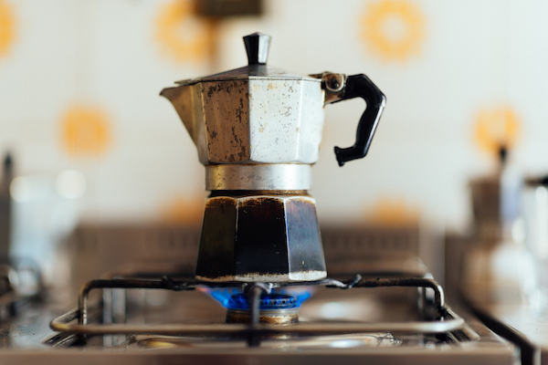 a metal coffee pot sits on a gas stove with a visible blue flame. the background features a blurry pattern of orange and white tiles.