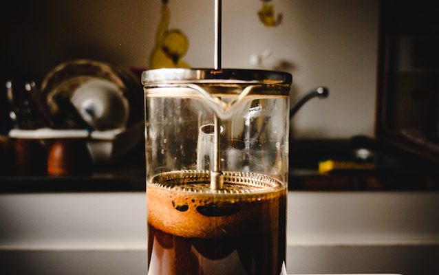 a French press full of coffee sits on a white surface in a dimly lit kitchen.