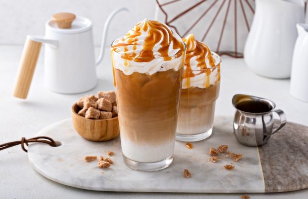 two iced caramel latte drinks topped with whipped cream and a drizzle of caramel, arranged on a marble countertop surrounded by a small pitcher, a bowl of brown sugar cubes and a white teapot.