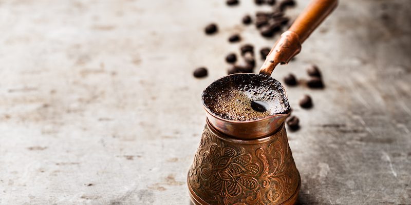 a copper cezve filled with freshly brewed coffee sits on a metal surface with coffee beans scattered in the background.