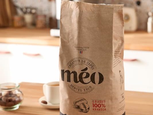 a bag of méo coffee with the words “excellence” is placed on the kitchen counter. next to it are two cups of coffee and in the background a carafe full of coffee beans.