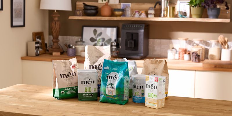 Five packs of méo brand coffee are displayed on a wooden kitchen counter, with shelves, a coffee machine and various kitchen items in the background.