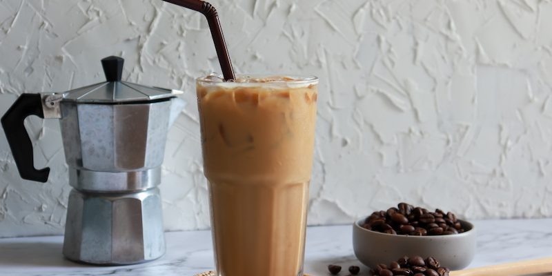 a glass of iced coffee with straw on a coaster, next to a bowl of coffee beans, a wooden spoon and a moka pot, all resting on a white surface.