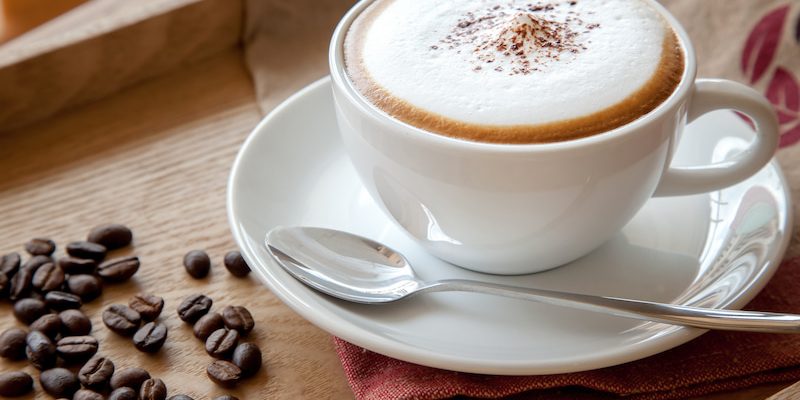 a cappuccino in a white cup on a saucer with a spoon, placed on a wooden tray. coffee beans are scattered nearby on a red cloth.