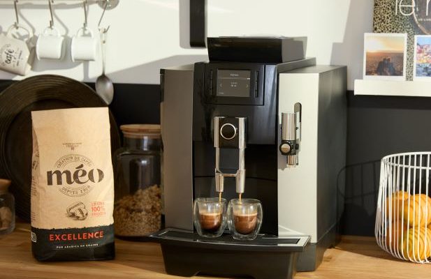 a modern espresso machine brews coffee in two transparent glasses on a wooden counter, next to a bag of coffee beans and a wire fruit basket with oranges. in the background there are shelves with cups.