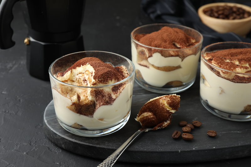 three glass cups containing tiramisu are placed on a slate tray. a spoon with tiramisu is placed in front, while in the background there is a mocha and coffee beans.