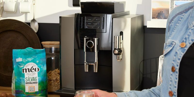 two people hold coffee cups in front of a silver automatic coffee machine sitting on the kitchen counter, with a bag of coffee beans nearby.