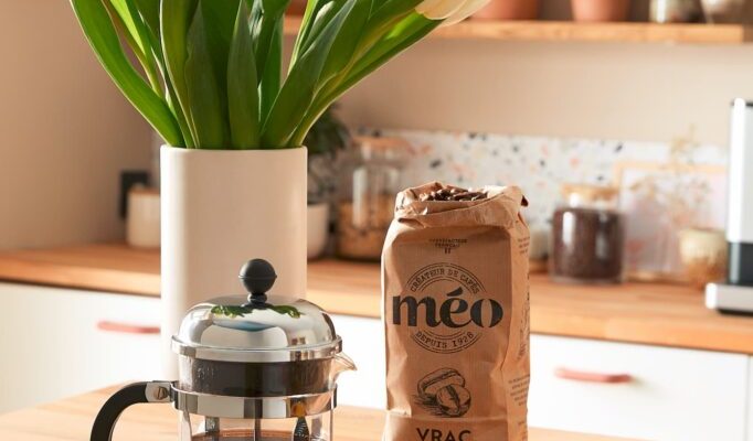 a French press coffee maker, a bag of coffee beans and a patterned mug are placed on a wooden counter next to a vase of white tulips in a bright, modern kitchen.