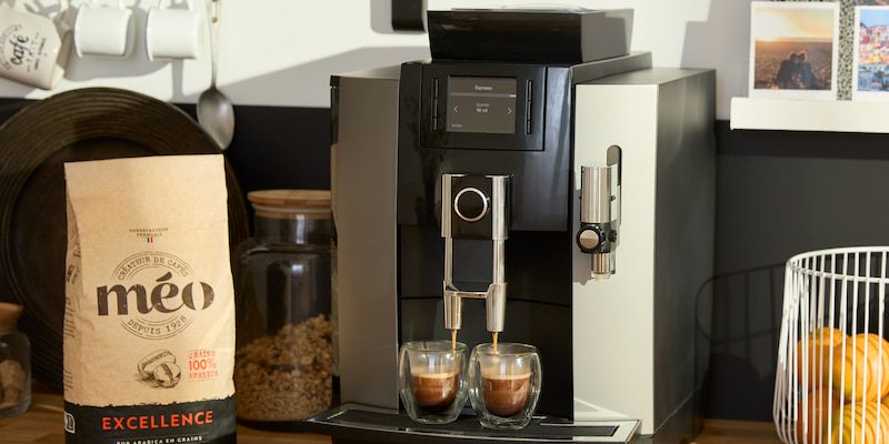 on the kitchen counter, a coffee machine prepares two espressos, next to a bag of meo coffee beans, a jar and a basket of oranges.