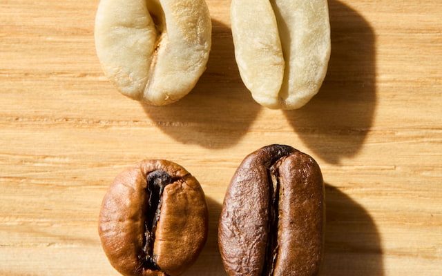 four coffee beans on a wooden surface; two are raw and light in color, and two are roasted and brown, illustrating the different stages of coffee processing.