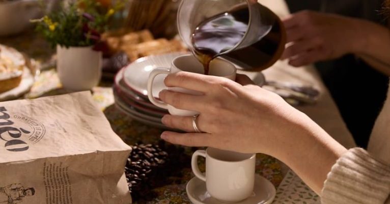 A person pours coffee from a glass carafe into a white cup placed on a table set with food, a bag of coffee beans and a potted plant.