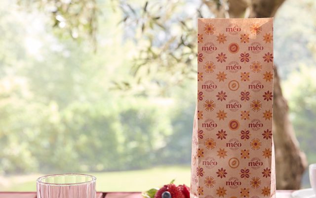 a glass of water, a labeled cup and saucer "distortion"fruit tarts and a patterned gift bag are placed on a red outdoor table with greenery in the background.